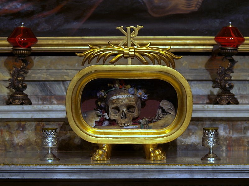 Golden container on a mantle holds skeletal remains with "St. Valentine" labeled across the forehead of the skull.