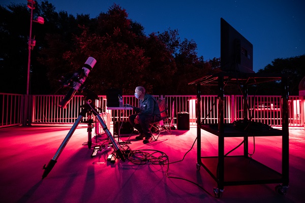 A man wearing a medical face mask sits at an outside computer and telescope setup at The George Observatory in the evening time.