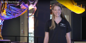 A smiling woman stands in front of a T. rex fossil display