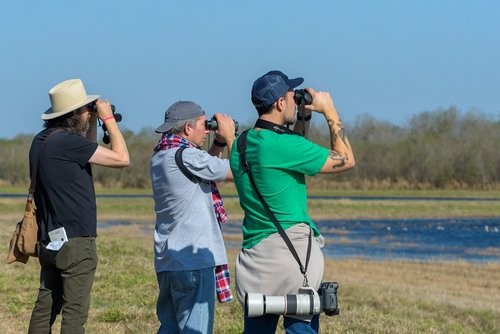 three men stand profile with binoculars looking out across a lake