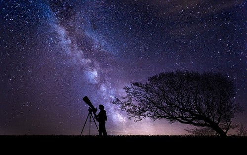 A person stands at a telescope underneath the stars